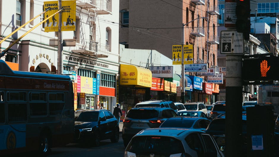 A busy urban street scene in Chinatown during daytime, featuring multiple parked and moving cars, including sedans and an SUV, in front of various storefronts with colourful signs in Chinese and English advertising books, collectibles, and other retail items. A pedestrian is seen walking along the sidewalk near a building with an entrance canopy. Overhead, there are yellow traffic signs and a traffic light displaying a red pedestrian signal. Visible features include a blue bus parked on the street with the label 'Clean Air Vehicle,' and a variety of vehicles are queued or moving through the area, demonstrating typical urban traffic during a home relocation or moving process. The environment is well-lit with natural sunlight, highlighting the diverse signage and building facades, which are part of the block where [COMPANY_NAME] provides removals and storage services, exemplifying the logistics involved in furniture transport and packing during house moves in Chinatown.