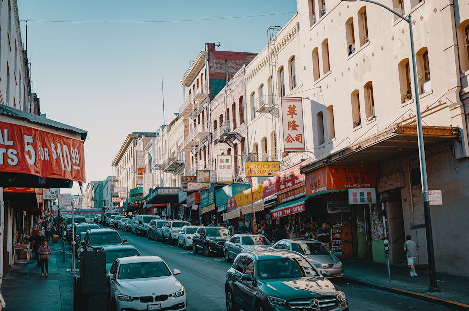 A street scene in Chinatown featuring a row of multi-storey buildings with various Asian signs and storefronts. Parked cars line both sides of the street, with some vehicles being used for furniture transport or home relocation purposes. The pavement is busy with pedestrians walking along storefronts and across the street. The buildings are a mix of white and brick facades, with awnings and shop signs in vibrant red, yellow, and green colors, indicating retail and dining establishments. Visible equipment includes a trolley used for loading items into a van, while the setting suggests a typical urban environment where home removals, packing, and moving logistics are carried out. The lighting indicates daytime, with natural sunlight illuminating the scene, and the overall atmosphere reflects a busy area where relocation services such as those offered by Man with Van Chinatown are applicable for moving home or furniture transport.