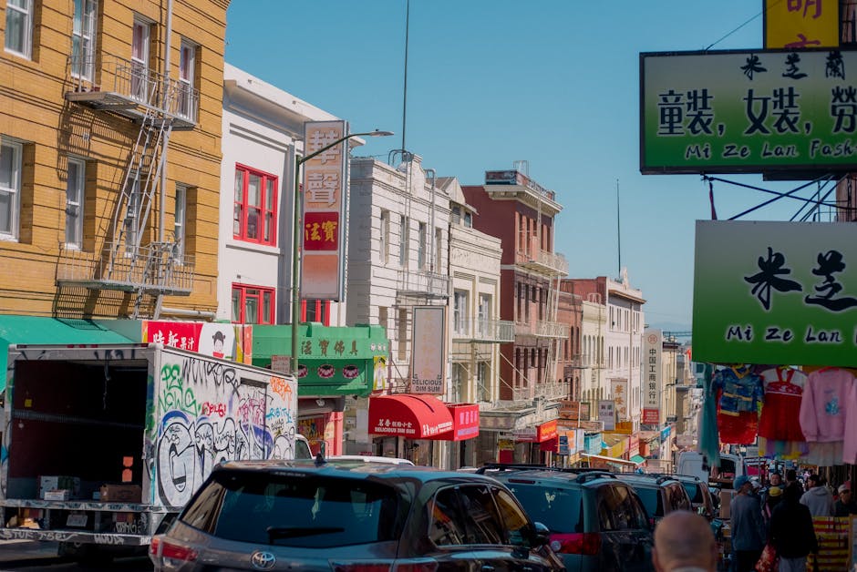 A busy urban street scene in Chinatown during daytime, featuring multiple parked and moving cars, including sedans and an SUV, in front of various storefronts with colourful signs in Chinese and English advertising books, collectibles, and other retail items. A pedestrian is seen walking along the sidewalk near a building with an entrance canopy. Overhead, there are yellow traffic signs and a traffic light displaying a red pedestrian signal. Visible features include a blue bus parked on the street with the label 'Clean Air Vehicle,' and a variety of vehicles are queued or moving through the area, demonstrating typical urban traffic during a home relocation or moving process. The environment is well-lit with natural sunlight, highlighting the diverse signage and building facades, which are part of the block where [COMPANY_NAME] provides removals and storage services, exemplifying the logistics involved in furniture transport and packing during house moves in Chinatown.