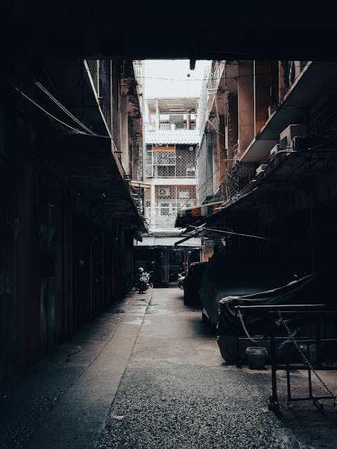 A narrow, urban alleyway during daytime with a view towards multi-storey residential buildings featuring exposed brick and metal balconies. The alley is lined with parked scooters, bicycles, and covered objects, possibly furniture or appliances, with some wrapped in protective plastic or fabric. The ground is paved with concrete and small gravel, and a metal chain barrier is visible on the right side, indicating restricted access. Overhead, there are wires and cables strung across between buildings, with a bright sky visible through the open space above. The scene depicts a residential exterior environment typical of city house removals, showing the tight access challenge faced during furniture transport or home relocation, and highlights the need for careful planning by [COMPANY_NAME], such as Man with Van Chinatown, when executing a house removal involving tight access and logistical coordination.