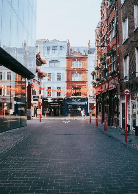 A street scene in Chinatown with a view of a narrow pedestrian walkway surrounded by multi-storey buildings with traditional brick facades, some featuring window flower boxes. The alley is lined with red bollards and string lights hanging across the street, creating a festive atmosphere. In the distance, there is a delivery van parked near the end of the alley, and the area appears to be part of a home relocation or furniture transport process managed by Man with Van Chinatown, as part of their house removals and moving services. The scene is captured during daylight with clear weather, emphasizing the urban environment typical of Chinatown's vibrant streets intertwined with commercial and residential buildings.