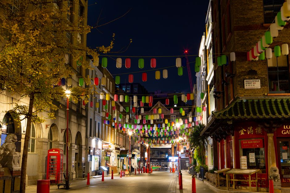 Nighttime street scene in Chinatown, featuring a row of multi-coloured lanterns hanging across the street between tall buildings with illuminated windows. The street is lined with commercial premises, including a Chinese restaurant with traditional architectural elements such as a green-tiled roof, and a red telephone box positioned near a tree with yellowing leaves. The pavement is partly blocked by orange traffic cones, and streetlights cast a warm glow on the scene. In the background, a van belonging to Man with Van Chinatown is being loaded with furniture and boxes wrapped in cardboard and plastic for a house relocation. The loading area shows furniture, moving boxes, and packing materials, with a focus on the logistics of furniture transport and packing for home removals and moving services within an urban environment.