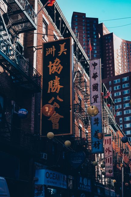Photograph of a busy urban street scene with tall, multi-storey brick buildings and a modern high-rise in the background. The buildings feature numerous vertical hanging signs with Chinese characters, illuminated by warm lighting, alongside decorative paper lanterns in various colors strung across the storefronts. Metal fire escape ladders and balconies are visible on the building facades. The street level shows shop fronts, and in the foreground, there is a pavement with some paraphernalia associated with moving processes, such as boxes, packing materials, or carts, suggesting a setting related to home relocation or furniture transport. The bright daylight enhances the lively atmosphere typical of Chinatown districts, aligning with the context of services like removals and moving logistics provided by companies such as Man with Van Chinatown.