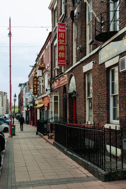 A busy urban street scene in Chinatown with multiple parked cars lining both sides of the road and a large white delivery truck actively unloading or loading alongside a row of storefronts. The storef