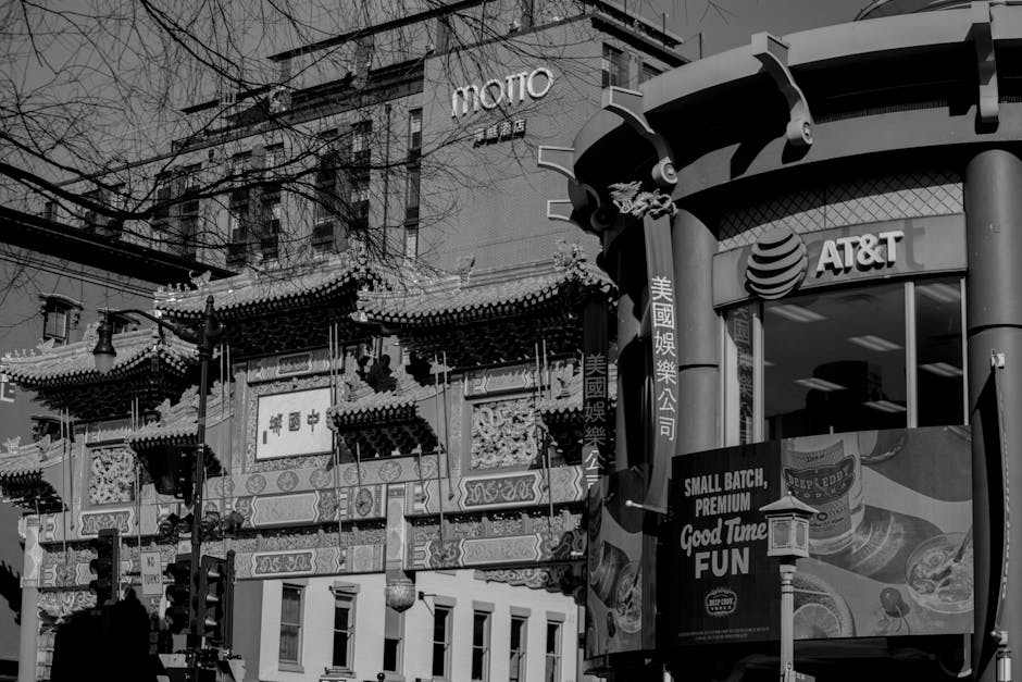 Black and white photograph showing a street scene in Chinatown with a traditional Chinese-style building featuring ornate roof detailing, decorative carvings, and Chinese characters on a signboard. In front of the building, a delivery van is parked, partially visible with its side door open, revealing packing materials such as cardboard boxes, plastic wrapping, and furniture blankets inside. A person, partially visible, is seen loading or unloading items onto the van using a trolley or dolly. The surrounding area includes commercial signage, with a modern high-rise building in the background displaying the word 'MOTTO' and an AT&T storefront with a curved glass facade. Overhead utility wires crisscross the sky, indicating an urban environment suited to a professional moving or home relocation service like Man with Van Chinatown, focused on furniture transport and packing during house moves.