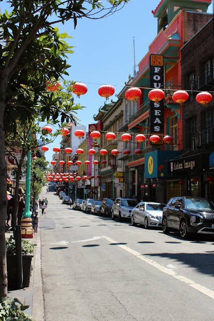 A street scene on Gerrard Street in Chinatown featuring a row of parked cars along the curb, with pedestrians walking on the sidewalk. The buildings on the right side display signs for various businesses, including a tofu eatery and a lounge, and are decorated with traditional red Chinese lanterns hanging overhead, creating a festive atmosphere. The street is lined with leafy trees providing partial shade, and the blue sky is visible above. This vibrant setting reflects the lively environment in which home relocation and furniture transport services by Man with Van Chinatown often take place, highlighting the urban context of moving logistics and loading areas for house removals in Chinatown.