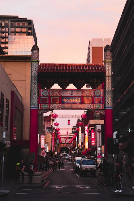 Black and white photograph showing a street scene in Chinatown with a traditional Chinese-style building featuring ornate roof detailing, decorative carvings, and Chinese characters on a signboard. In front of the building, a delivery van is parked, partially visible with its side door open, revealing packing materials such as cardboard boxes, plastic wrapping, and furniture blankets inside. A person, partially visible, is seen loading or unloading items onto the van using a trolley or dolly. The surrounding area includes commercial signage, with a modern high-rise building in the background displaying the word 'MOTTO' and an AT&T storefront with a curved glass facade. Overhead utility wires crisscross the sky, indicating an urban environment suited to a professional moving or home relocation service like Man with Van Chinatown, focused on furniture transport and packing during house moves.