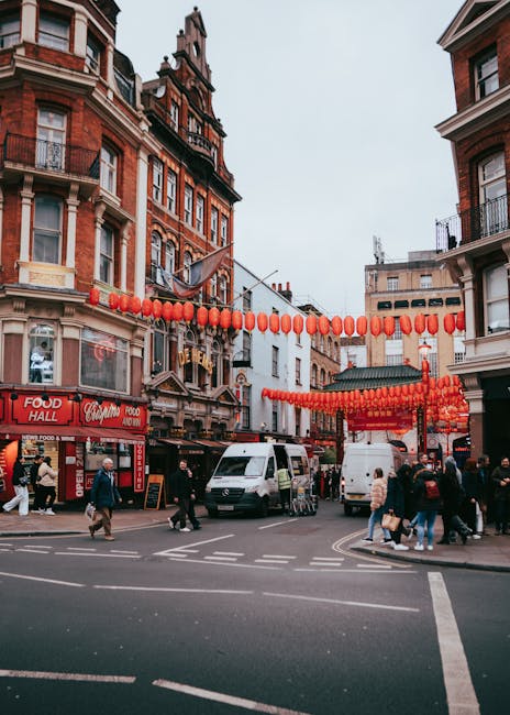A busy street scene in Leicester Square Chinatown during daytime, with multiple pedestrians crossing at a marked crosswalk and walking along the pavement. The street is lined with historic red-brick buildings featuring decorative architectural details, and red paper lanterns are strung overhead across the street, creating a festive atmosphere. Several vehicles, including a white van and a small delivery truck, are parked or moving through the intersection, indicative of a home relocation or furniture transport process. The foreground shows a loading area with stacked cardboard boxes, wrapped furniture, and packing materials placed on the sidewalk near the vehicles. The scene suggests active packing and loading activities associated with moving services provided by Man with Van Chinatown, with a focus on efficient furniture transport and house removals within the vibrant Chinatown district.