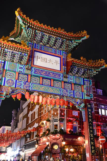 Nighttime view of a traditional ornate Chinese gate in Leicester Square Chinatown, decorated with colorful intricate patterns, dragon motifs, and bright lanterns hanging along the street. In the foreground, there are several cardboard boxes and packing materials placed on the pavement, alongside a large moving trolley partially visible, indicating an ongoing home relocation process. The scene suggests a busy environment where furniture, household items, and packing supplies are being prepared for transportation. The illuminated signage of Man with Van Chinatown can be seen on a van parked at the curb, reflecting typical logistics involved in house removals and furniture transport within the city. The vibrant lighting highlights the detailed craftsmanship of the arch and the bustling atmosphere of the Chinatown district at night, emphasizing the importance of careful packing and loading procedures in professional removals.