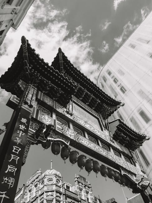 A busy street scene in Leicester Square Chinatown during daytime, with multiple pedestrians crossing at a marked crosswalk and walking along the pavement. The street is lined with historic red-brick buildings featuring decorative architectural details, and red paper lanterns are strung overhead across the street, creating a festive atmosphere. Several vehicles, including a white van and a small delivery truck, are parked or moving through the intersection, indicative of a home relocation or furniture transport process. The foreground shows a loading area with stacked cardboard boxes, wrapped furniture, and packing materials placed on the sidewalk near the vehicles. The scene suggests active packing and loading activities associated with moving services provided by Man with Van Chinatown, with a focus on efficient furniture transport and house removals within the vibrant Chinatown district.