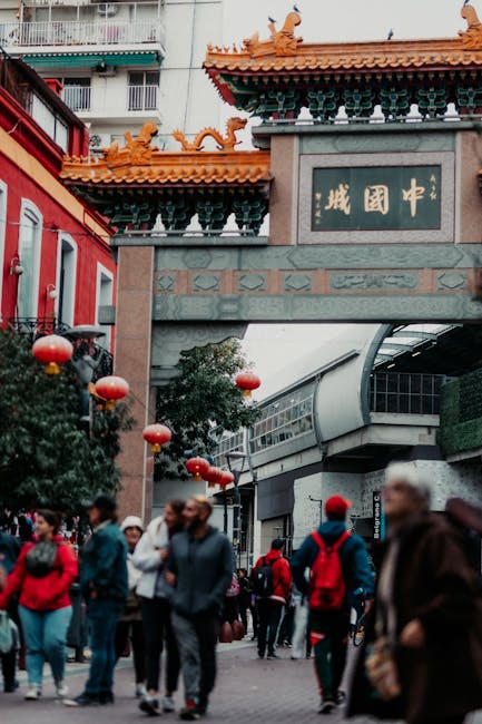 The image shows a busy outdoor scene in Chinatown with numerous pedestrians walking beneath a traditional Chinese-style gate adorned with decorative dragons and Chinese characters. Red lanterns hang from greenery and buildings on either side, contributing to the festive atmosphere. In the foreground, a group of people in casual clothing and backpacks are walking on the pavement, some engaged in conversation. Inside a nearby commercial building, visible through large glass windows, there are cardboard boxes, plastic wrapping materials, and possibly some furniture being prepared for transport or home relocation. A loading area adjacent to the building shows a vehicle or van partially visible, suggesting ongoing furniture transport or packing activity as part of house removals services. The overall scene captures a vibrant, culturally themed urban environment typical of Chinatown, with elements related to moving logistics subtly integrated into the context of packing and furniture transport facilitated by [COMPANY_NAME].