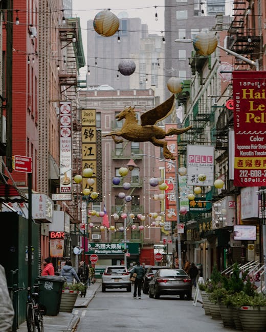 A busy urban street scene in Chinatown, featuring a narrow roadway with several parked and moving cars, flanked by tall buildings with numerous colourful signs in Chinese characters advertising various shops, restaurants, and services. Overhead, decorative paper lanterns and a large, realistic model of a flying dragon are suspended above the street, creating a festive atmosphere. Pedestrians are seen walking on the sidewalks, some near the storefronts, while others are entering or exiting buildings. On the left side, a green waste bin and bicycles are visible, with retail and commercial establishments including a bar and a hair salon on both sides of the street. The scene is captured during daylight with diffused lighting, highlighting the details of the street life and decorations, which support the context of a lively area often involved in home and business relocation activities, as handled by Man with Van Chinatown's removals services.