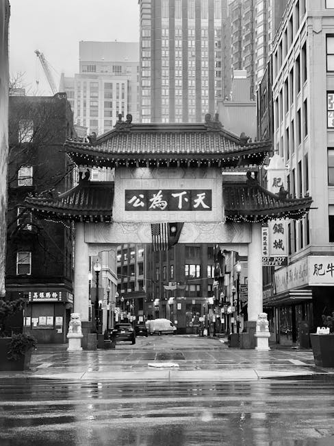 The image shows a busy outdoor scene in Chinatown with numerous pedestrians walking beneath a traditional Chinese-style gate adorned with decorative dragons and Chinese characters. Red lanterns hang from greenery and buildings on either side, contributing to the festive atmosphere. In the foreground, a group of people in casual clothing and backpacks are walking on the pavement, some engaged in conversation. Inside a nearby commercial building, visible through large glass windows, there are cardboard boxes, plastic wrapping materials, and possibly some furniture being prepared for transport or home relocation. A loading area adjacent to the building shows a vehicle or van partially visible, suggesting ongoing furniture transport or packing activity as part of house removals services. The overall scene captures a vibrant, culturally themed urban environment typical of Chinatown, with elements related to moving logistics subtly integrated into the context of packing and furniture transport facilitated by [COMPANY_NAME].