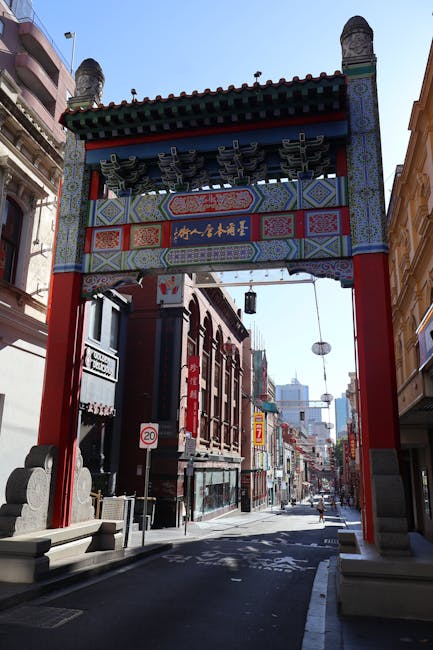 A large, traditional Chinese-style archway with red columns, decorative green and blue roof tiles, and intricate patterns in red, gold, and blue, spans across a city street in Chinatown. The arch features Chinese characters on a horizontal plaque in gold against a dark background and is supported by stone bases. Beyond the arch, commercial buildings with brick facades and various signage line the street, with some signs indicating businesses and speed limits visible. The street is relatively empty, with no visible pedestrians or vehicles. The sky above is clear, and a few modern high-rise buildings are seen in the background. The scene suggests a typical urban environment where the arch serves as an entrance to Chinatown, with a focus on the cultural landmark amidst city traffic and buildings, aligning with house removals or relocation contexts involving access and transport solutions managed by Man with Van Chinatown.