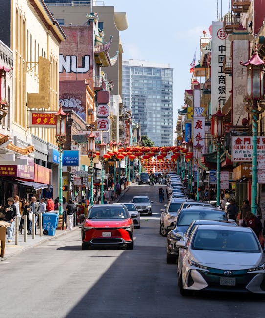 A large, traditional Chinese-style archway with red columns, decorative green and blue roof tiles, and intricate patterns in red, gold, and blue, spans across a city street in Chinatown. The arch features Chinese characters on a horizontal plaque in gold against a dark background and is supported by stone bases. Beyond the arch, commercial buildings with brick facades and various signage line the street, with some signs indicating businesses and speed limits visible. The street is relatively empty, with no visible pedestrians or vehicles. The sky above is clear, and a few modern high-rise buildings are seen in the background. The scene suggests a typical urban environment where the arch serves as an entrance to Chinatown, with a focus on the cultural landmark amidst city traffic and buildings, aligning with house removals or relocation contexts involving access and transport solutions managed by Man with Van Chinatown.