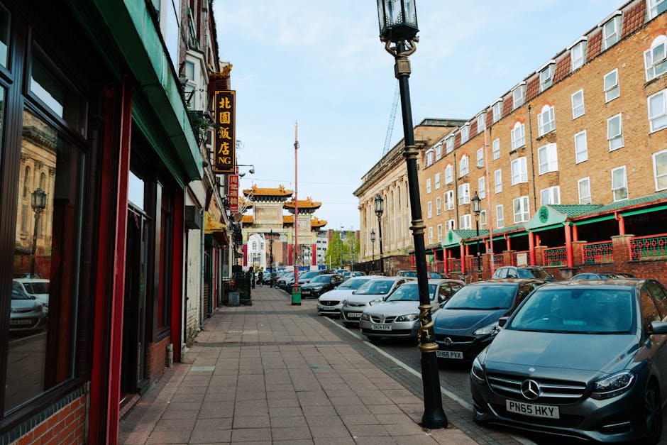 A street scene in Chinatown with a traditional Chinese gate at the end of the road, decorated with red lanterns and intricate architectural details. Parked cars line both sides of the street, and pedestrians walk along the pavement. In the foreground, a man from Man with Van Chinatown is seen loading or unloading several cardboard boxes and wrapped furniture onto or from a van positioned partially on the pavement. The scene takes place during daylight with overcast skies, and surrounding buildings include a mix of older brick structures and modern glass facades. This image illustrates the process of house removals, highlighting the packing and furniture transport involved in home relocation services offered by Man with Van Chinatown, demonstrating their efficient approach to moving objects within and out of Chinatown flats.