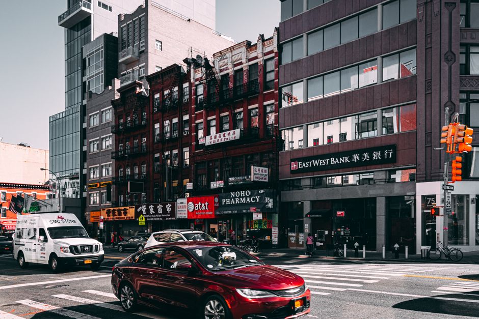 A busy urban street scene in Chinatown near WC2H, showing several multi-storey commercial and office buildings with modern glass facades and traditional brick facades. On the ground level, there are s