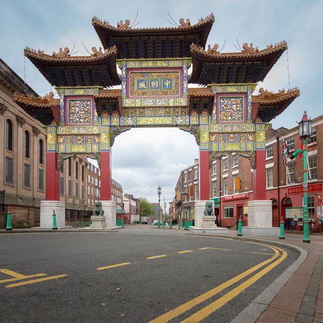 A large, ornate Chinese-style archway with intricate colorful patterns and traditional roof decorations spans across an empty city street, flanked by historic brick buildings. The archway features Chinese characters on a central plaque, supported by four red columns, each topped with decorative elements. Stone lion statues are positioned at the base of the columns, serving as guardians. The street beneath is paved with dark asphalt, marked by double yellow lines, and appears to be closed off to traffic, possibly for a relocation or event. On the right side, a storefront with signage and a Chinese lantern decoration is visible, indicating a Chinatown district. The lighting suggests daytime with a cloudy sky, and there are no pedestrians or vehicles present, suggesting a quiet moment during packaging and moving preparations. This scene reflects typical elements involved in house removals or interior packing in a Chinatown area, supported by the services of [COMPANY_NAME].