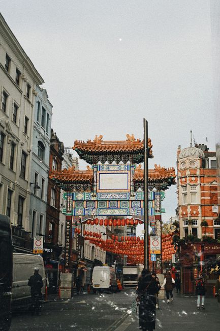 A street scene in Chinatown featuring a large, ornate Chinese-style gateway with intricate blue, red, and gold decorative elements, including traditional curved roof tiles and hanging red lanterns. Surrounding the gateway are multi-story buildings with classic architectural details, and pedestrians walking along the pavement. In the foreground, a man is loading or unloading cardboard boxes and plastic-wrapped furniture onto or from a commercial van parked near the curb. The van is positioned partly inside a loading area adjacent to the building, with some furniture covered in blankets and secured with straps. The scene is set during daylight under a clear sky, with a few street lamps and signage visible, indicating a busy urban environment. This image illustrates the logistics involved in home relocation and furniture transport within the Chinatown district, supported by Man with Van Chinatown’s removal services, including packing, moving, and loading processes connected to Chinatown removals and permit management for street access.