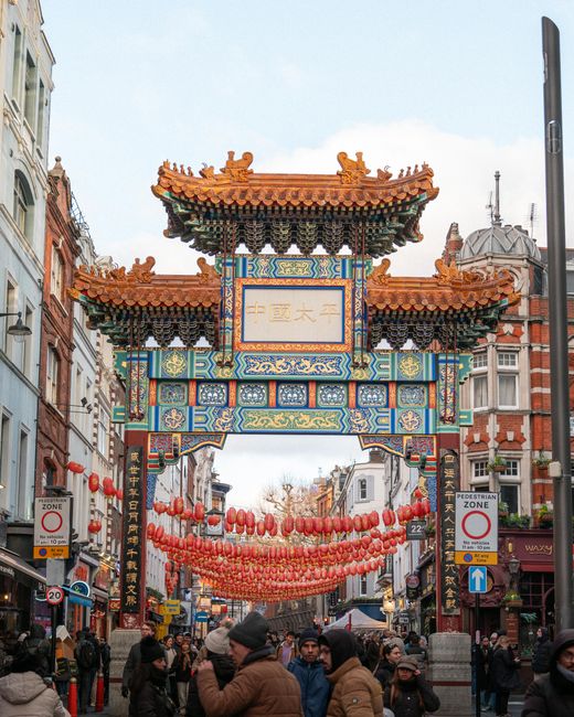 A large, ornate Chinese-style archway with intricate colorful patterns and traditional roof decorations spans across an empty city street, flanked by historic brick buildings. The archway features Chinese characters on a central plaque, supported by four red columns, each topped with decorative elements. Stone lion statues are positioned at the base of the columns, serving as guardians. The street beneath is paved with dark asphalt, marked by double yellow lines, and appears to be closed off to traffic, possibly for a relocation or event. On the right side, a storefront with signage and a Chinese lantern decoration is visible, indicating a Chinatown district. The lighting suggests daytime with a cloudy sky, and there are no pedestrians or vehicles present, suggesting a quiet moment during packaging and moving preparations. This scene reflects typical elements involved in house removals or interior packing in a Chinatown area, supported by the services of [COMPANY_NAME].